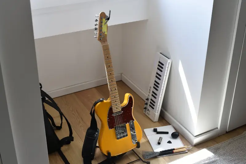 Guitare électrique à corps plein jaune posée sur son stand, sangle, médiator et bottleneck au sol près d'un petit clavier