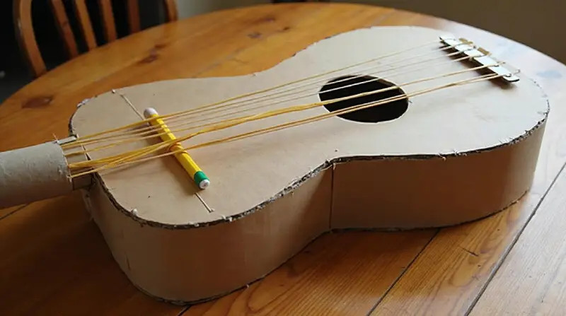 Caisse en carton découpée en forme de guitare, élastiques tendus au-dessus d'un trou rond, crayon jaune glissé dessous, manche en rouleau de carton