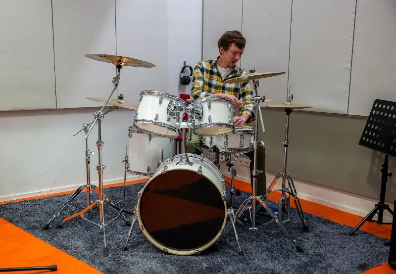 Un batteur assis à une batterie acoustique blanche posée sur un tapis, dans une salle de répétition