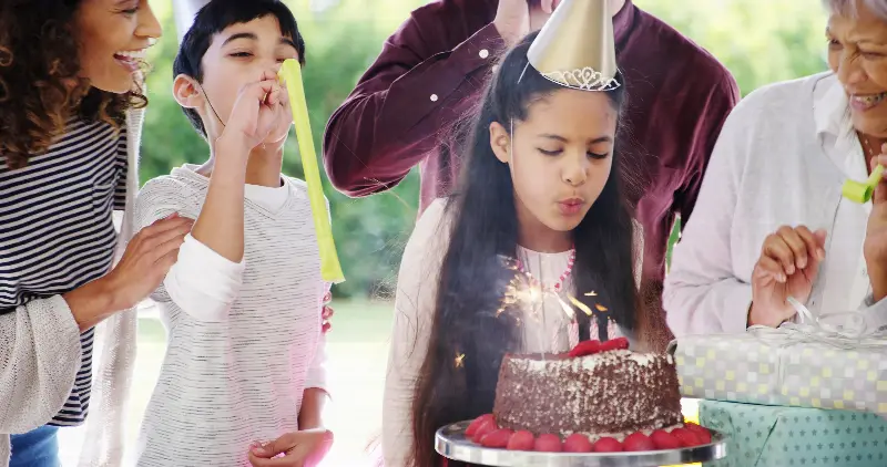 Fillette soufflant les bougies de son gâteau d'anniversaire, entourée de sa famille avec chapeaux pointus et langues de belle-mère