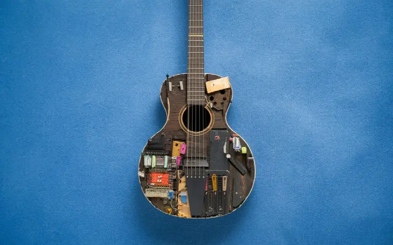 Guitare acoustique dont la table est recouverte de pièces de récupération, composants électroniques et morceaux de bois, posée sur un fond bleu