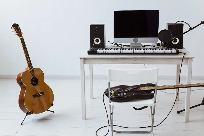 Guitare acoustique sur son stand, basse électrique posée sur une chaise et clavier maître devant un ordinateur, dans un petit studio à la maison