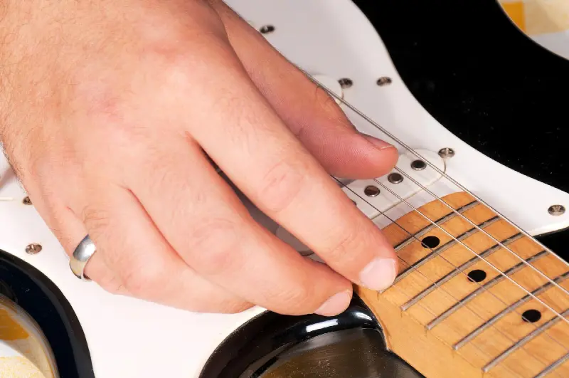 Doigt de la main droite posé sur les cordes, au bout du manche d'une guitare électrique noire et blanche
