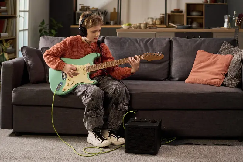 Jeune guitariste assis sur un canapé, casque sur les oreilles, guitare branchée sur un petit ampli posé au sol