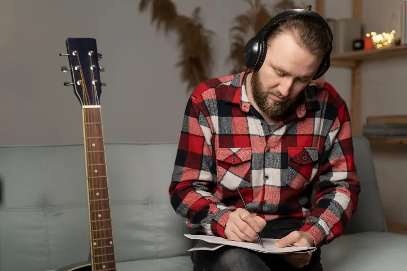 Guitariste casque sur les oreilles qui note dans un cahier, une guitare posée à côté de lui sur le canapé