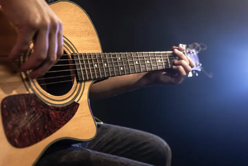 Guitariste assis jouant une guitare folk à pan coupé, main gauche formant un accord, sur fond sombre éclairé en bleu