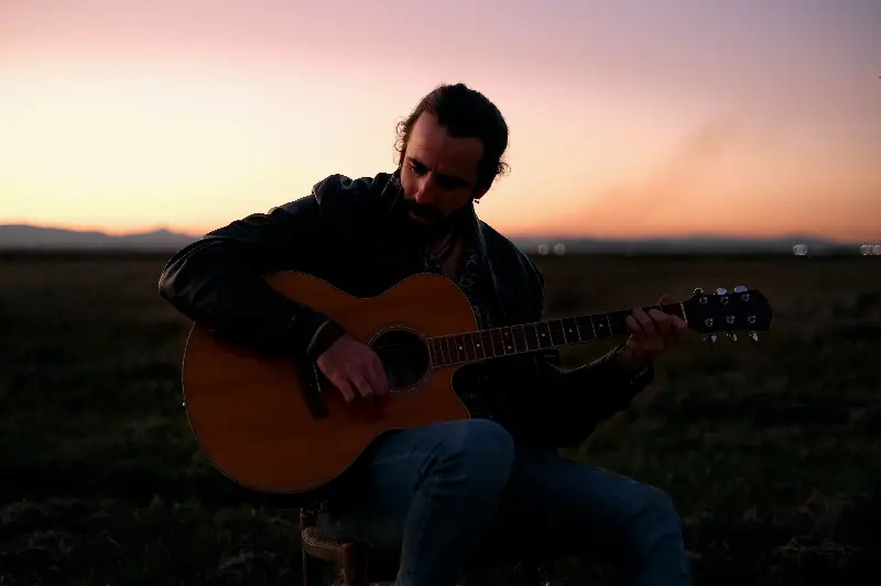 Guitariste barbu assis dans une plaine au crépuscule, jouant d'une guitare acoustique