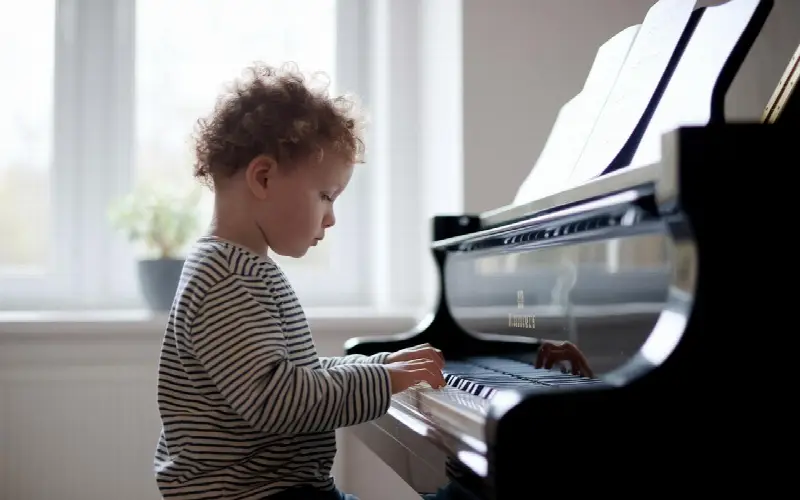 Jeune enfant en pull rayé, les deux mains posées sur le clavier d'un piano à queue noir, près d'une fenêtre