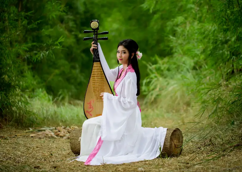 Jeune femme en robe traditionnelle blanche et rose, assise sur un tronc dans un bois de bambous, tenant un pipa à la verticale