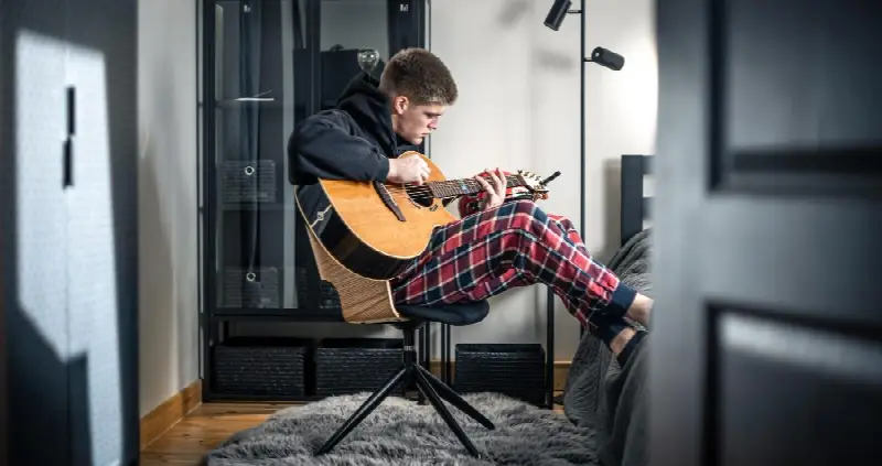 Jeune homme en pyjama à carreaux jouant d'une guitare folk à pan coupé, assis sur un tabouret dans sa chambre