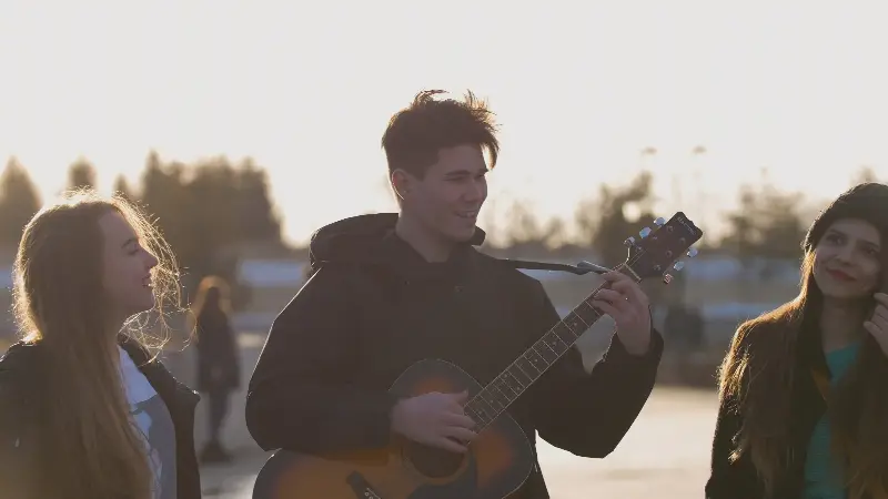 Jeune homme qui joue de la guitare acoustique en plein air au coucher du soleil, deux amies l'écoutent en souriant