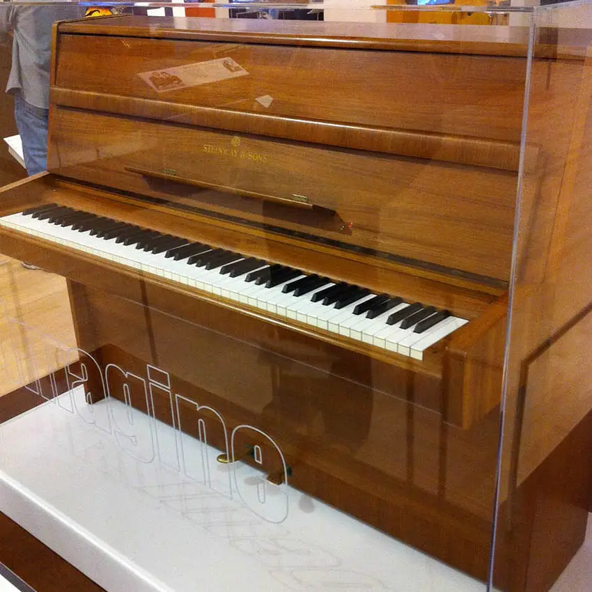 Piano droit Steinway en noyer verni exposé sous une vitrine, le mot Imagine inscrit en blanc sur la paroi transparente