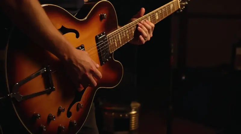 Mains d'un guitariste sur une guitare jazz à caisse creuse, ouïes en f et vernis sunburst, dans une salle sombre