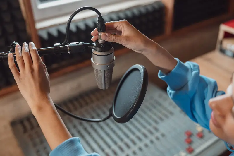 Mains réglant un micro de studio derrière son filtre anti-pop, au-dessus d’une console de mixage