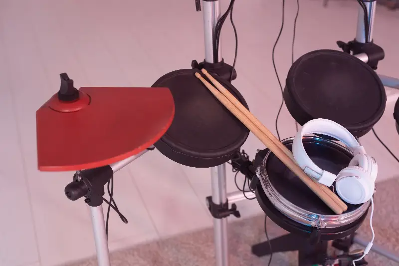 Pads noirs d'une batterie électronique sur leur rack, cymbale rouge, baguettes et casque blanc posés sur la caisse claire