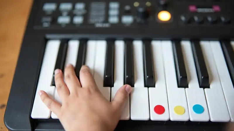 Petite main d'enfant posée sur les touches blanches d'un clavier électronique, trois touches marquées de gommettes rouge, jaune et bleue