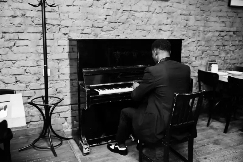 Pianiste en costume vu de dos, assis à un piano droit contre un mur de briques, en noir et blanc