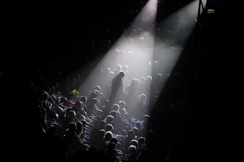 Public assis dans les gradins d'une salle de spectacle, traversé par deux faisceaux de projecteurs dans la pénombre