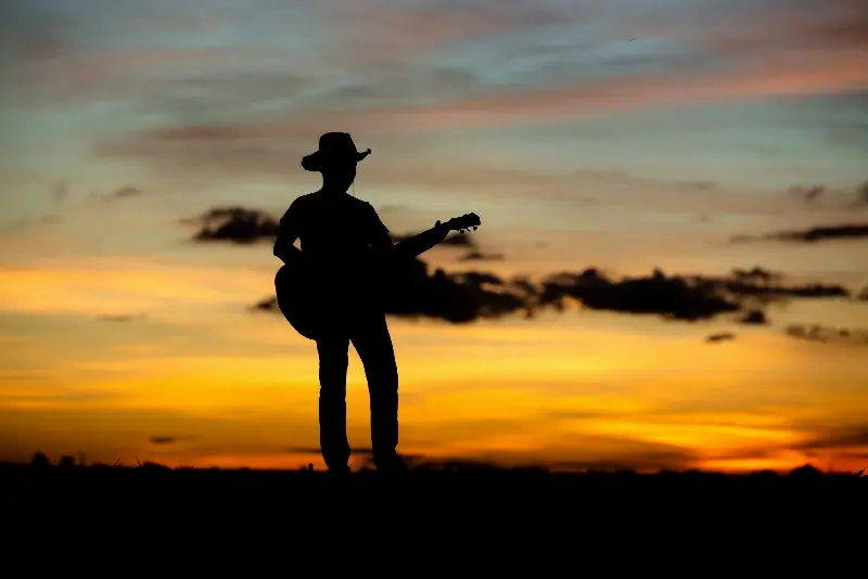 Silhouette d'un homme en chapeau de cow-boy jouant de la guitare acoustique dans un champ, au coucher du soleil