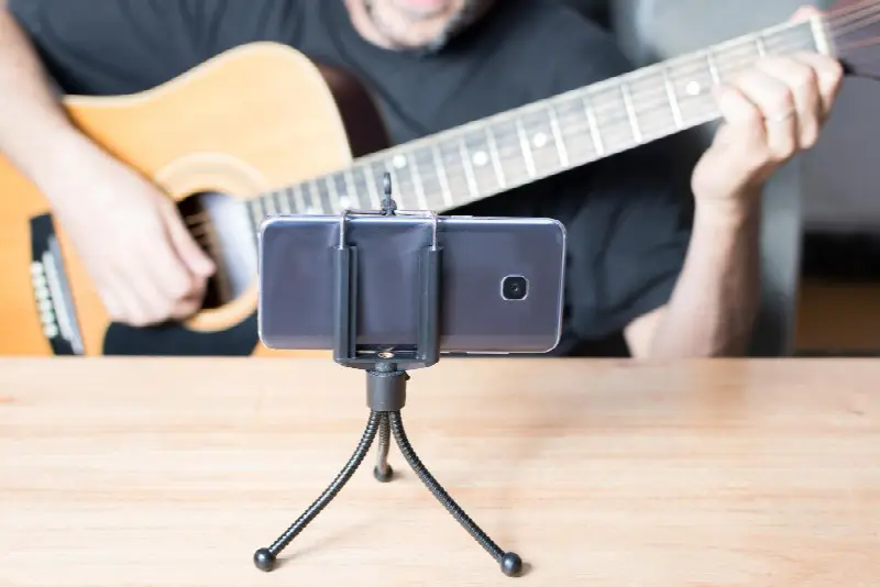Smartphone posé sur un petit trépied devant un homme qui joue d'une guitare folk
