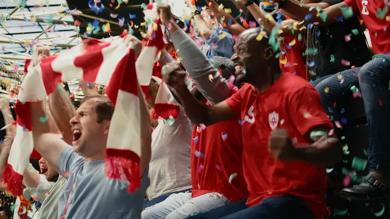 Supporters en maillots rouges qui chantent dans une tribune, écharpes levées, sous une pluie de confettis