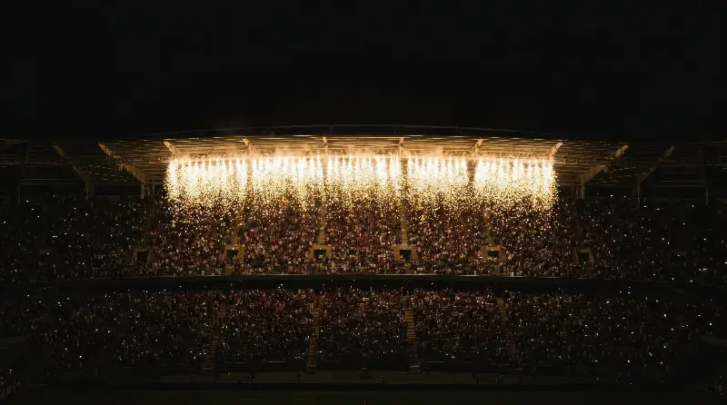 Tribunes d'un stade plein, de nuit, sous des gerbes d'étincelles qui tombent du toit