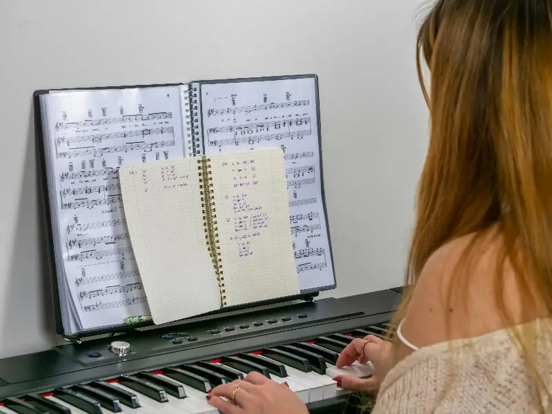 Une pianiste joue sur un piano numérique, un carnet d'accords notés à la main ouvert devant une partition