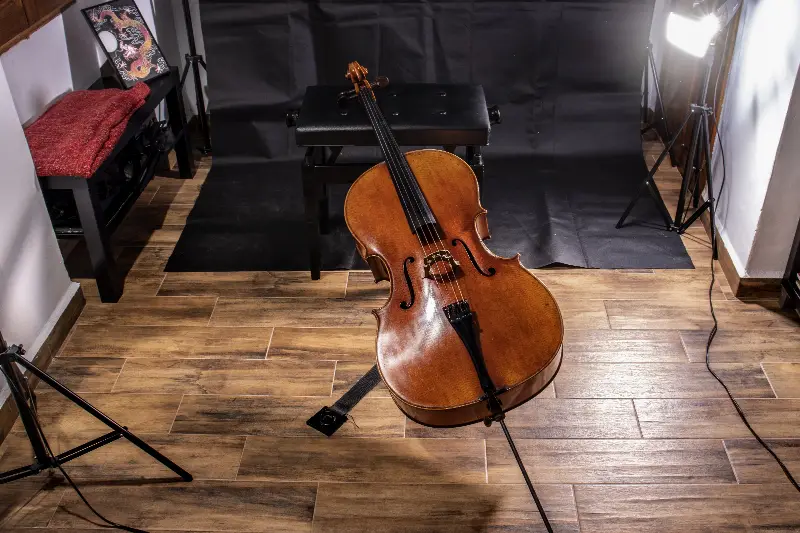 Violoncelle posé au sol devant un banc, dans un petit studio éclairé par des projecteurs